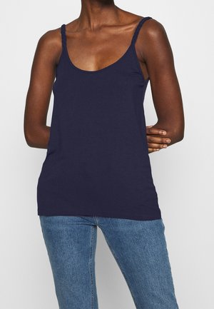 Woman wearing a navy blue tank top with braided straps and light blue denim jeans, standing with one arm across her waist.