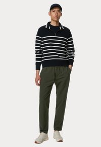 Felpa a zip a righe navy e bianche, pantaloni verde oliva con cordino e scarpe da ginnastica chiare indossati da un modello. Design semplice e moderno con texture morbide.