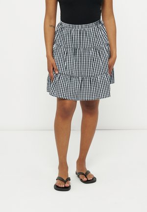 Kobieta ubrana w czarno-białą, warstwową spódnicę w kratę, czarny bezrękawnik oraz czarne klapki, stojąca na białym tle.