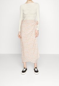 Un haut crème à manches longues avec un détail froncé, associé à une jupe rose ornée de petites fleurs oranges, le tout porté avec des baskets noires.