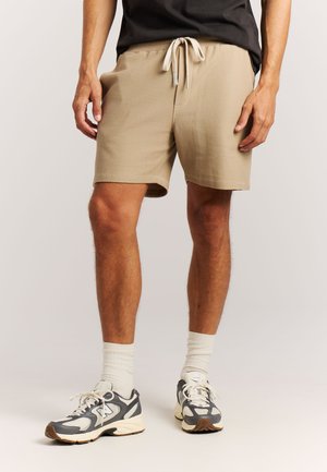 Mann trägt beige Shorts mit Kordelzug, weiße Sportsocken und graue und weiße Sneaker, steht vor einem neutralen Hintergrund.