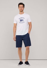 Camiseta blanca con estampado gráfico en azul marino, combinada con pantalones cortos azul marino. El modelo lleva zapatillas oscuras. Diseño simple, ajuste cómodo, estilo casual.