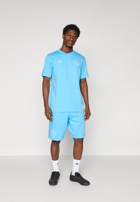 Light blue cotton t-shirt and shorts, featuring white logo accents. Model wears black shoes and white socks. Smooth texture, relaxed fit.