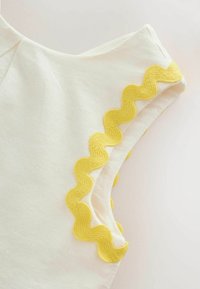 Gros plan d’un tissu blanc avec un galon ondulé jaune le long du bord de l’emmanchure d’un vêtement sans manches.