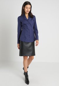 Marineblaue Bluse mit langen Ärmeln, mit geknöpftem Kragen, Frontknopfverschluss und taillierter Form, kombiniert mit einem schwarzen Leder-Bleistiftrock.
