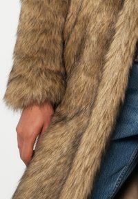 Καφέ τεχνητό γούνες παλτό με μαλακή, μακριά υφή. Εντυπωσιακή γυαλάδα και σχεδιασμένα νήματα. Βραχίονας φαίνεται να κρατάει το ύφασμα ενάντια σε τζιν.