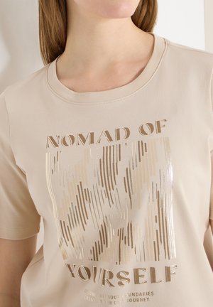Frau trägt ein beiges T-Shirt mit goldenem vertikalem Liniendesign und dem Text "Nomad of Yourself" sowie einem kleineren Slogan darunter.
