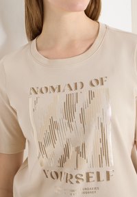 Femme portant un t-shirt beige avec un design de ligne verticale dorée et le texte « Nomade de soi-même » suivi d'un slogan plus petit en dessous.