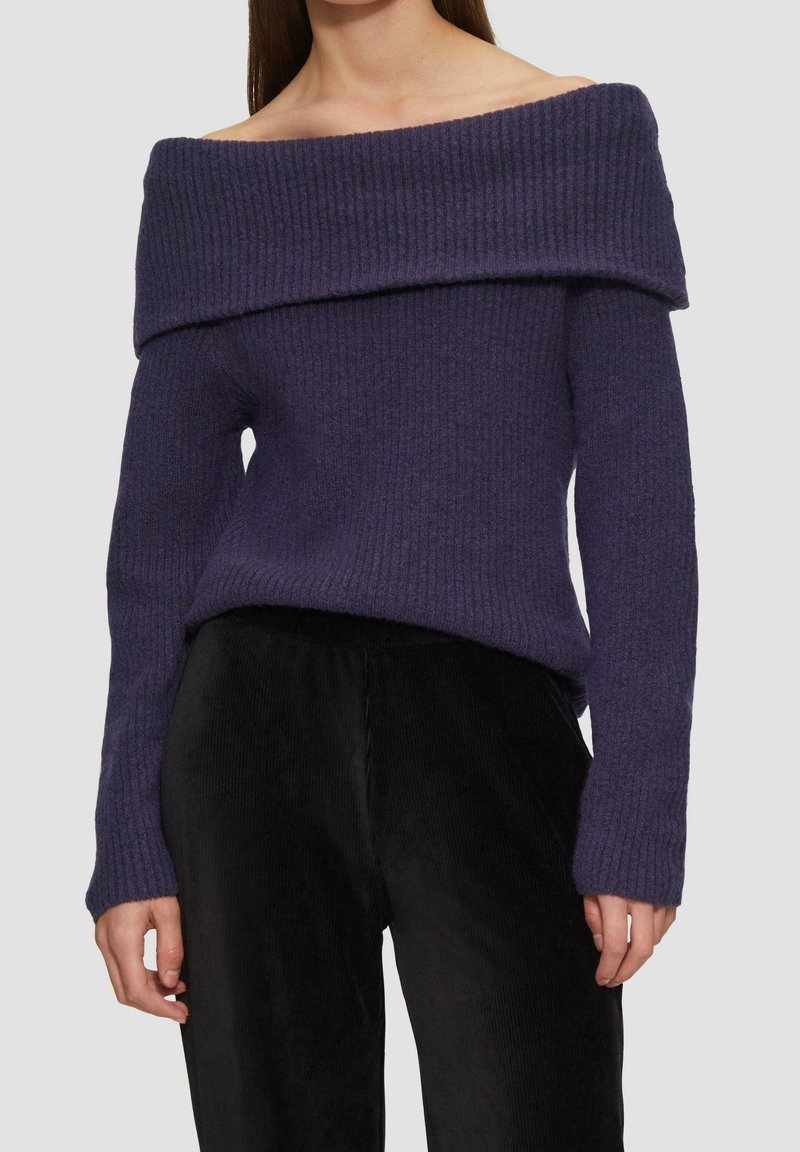 Femme portant un pull à manches tombantes en tricot côtelé violet foncé et un pantalon noir texturé sur un fond uni.