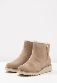 Botas de tobillo de ante beige con punta redondeada, forro de piel suave en la parte superior y cremalleras laterales con acentos de metal dorado.