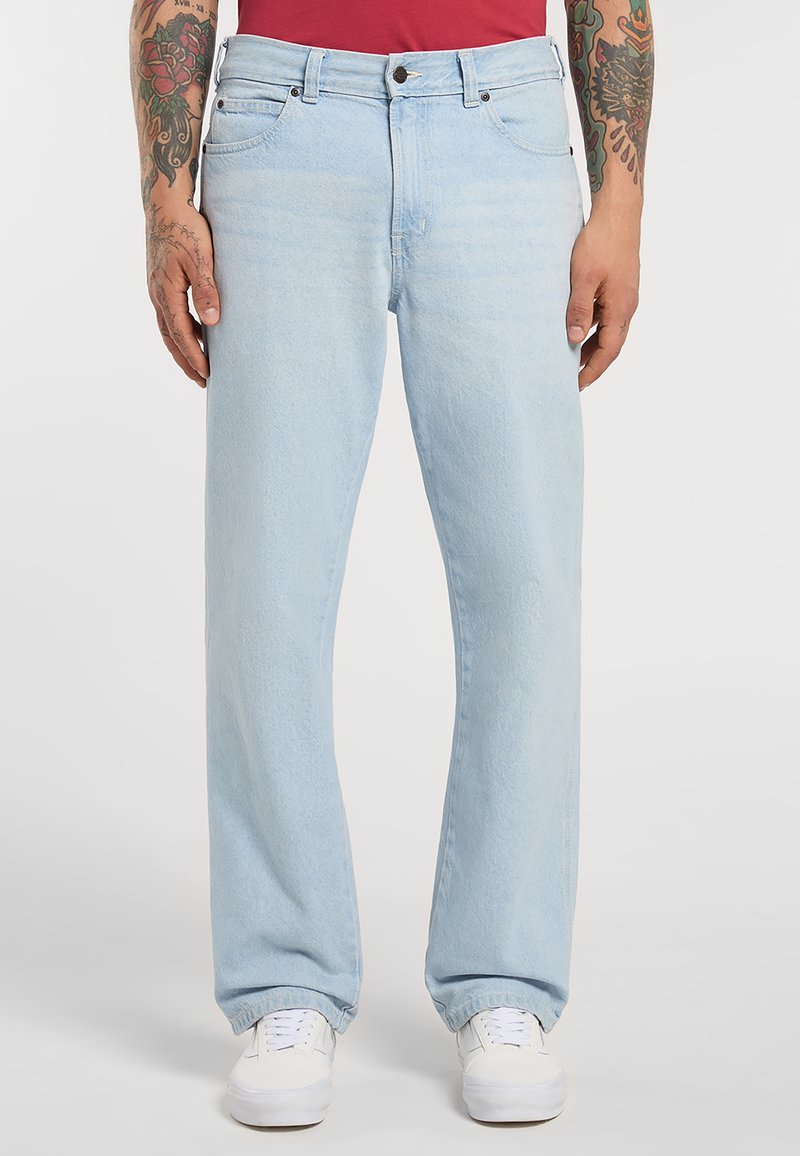 Lichtblauwe jeans met rechte pijpen, gedragen door een getatoeëerde persoon in witte sneakers en een rood shirt, staand tegen een witte achtergrond.