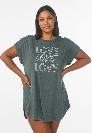 Kvinde med mellem-lang mørkt hår iført en løs grøn kjole med ordet "LOVE" trykt tre gange i hvid tekst.