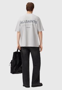 T-shirt oversize gris clair avec le logo "ALLSAINTS" en gris foncé au dos, associé à un jean large noir et un sac noir.