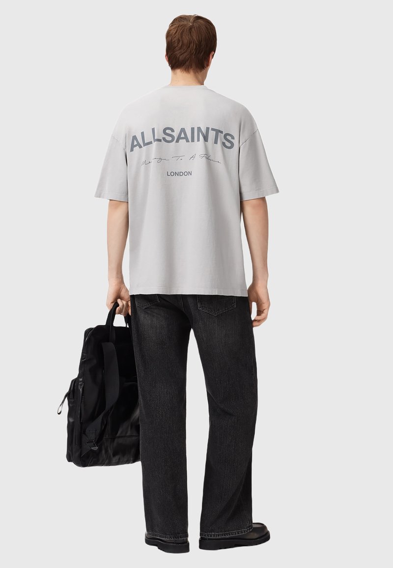 T-shirt oversize gris clair avec le logo "ALLSAINTS" en gris foncé au dos, associé à un jean large noir et un sac noir.