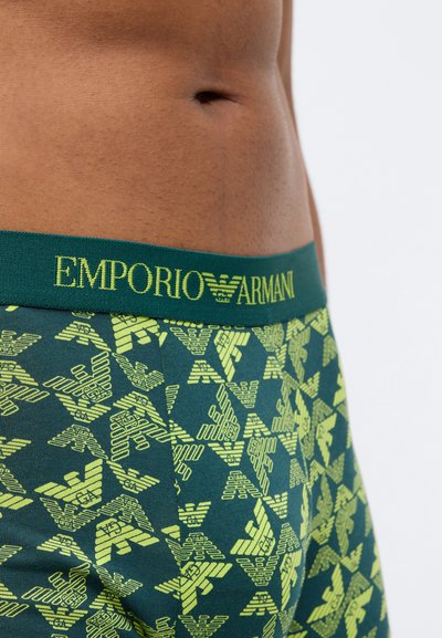 Žali bokseriai su geltonu geometriniu raštu visame paviršiuje. Elastinė juosta su kontrastinga geltona spalva turi užrašą "EMPORIO ARMANI".