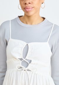 Vestido blanco con un corte en forma de llave, atado a la cintura, usado sobre una blusa de manga larga de canalé gris claro. Tela suave, detalles delicados.