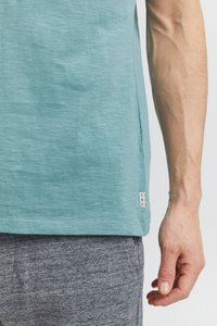 Svetlomodrá košeľa s krátkym rukávom vyrobená z mäkkej, textúrovanej látky. Na spodnej ľavej strane je viditeľná malá značka. Sivé šortky sú čiastočne zobrazené.