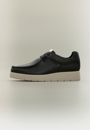 Chaussure décontractée en cuir noir texturé avec des lacets noirs et une semelle épaisse en caoutchouc beige clair, vue de profil sur un fond neutre.