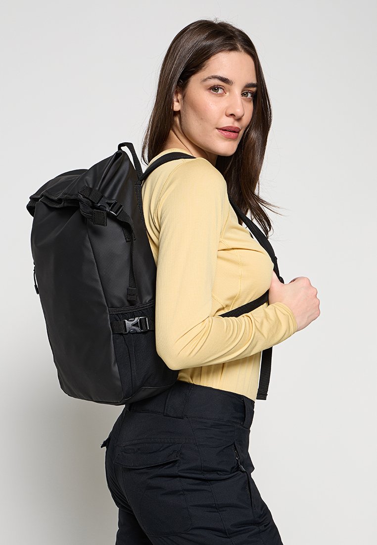 Czarny plecak z klapką, z regulowanymi paskami, masywnymi metalowymi elementami i teksturowaną powierzchnią, noszony na jednym ramieniu, na żółtej koszuli.
