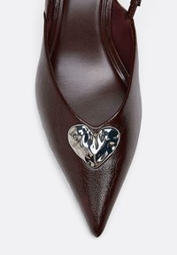 Brauner Lederpumps mit spitzer Vorderkappe und glänzendem silbernen Herzdetail auf dem Vorderblatt. Verfügt über ein Slingback-Design für zusätzlichen Stil.