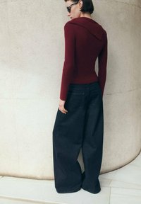 Maglione bordeaux a coste con un ampio colletto abbinato a pantaloni di jeans scuri e ampi. L'outfit presenta una superficie testurizzata e una silhouette rilassata.