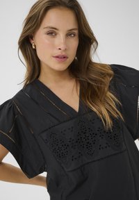 Fekete blúz V-kivágással, hímzett csipkés díszítéssel és nyitott mintázattal, rövid puffos ujjakra, sima textúrával.