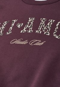 Tissu marron avec le texte "MI AMOR" en imprimé léopard et "Studio Club" en écriture cursive beige en dessous sur un vêtement avec encolure.