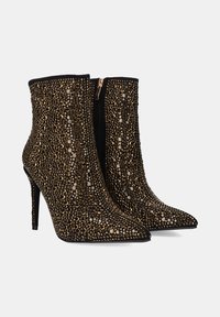 Botas de tobillo negras con puntas afiladas, adornadas con tachuelas metálicas en tonos dorados, tacones altos de aguja y un cierre con cremallera lateral.