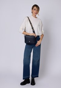 Námorně modrá kožená crossbody taška s texturovaným povrchem a vzorovaným popruhem. Modelka má na sobě bílé svetr a modré volné džíny. Černé boty.