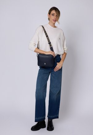 Námorně modrá kožená crossbody taška s texturovaným povrchem a vzorovaným popruhem. Modelka má na sobě bílé svetr a modré volné džíny. Černé boty.