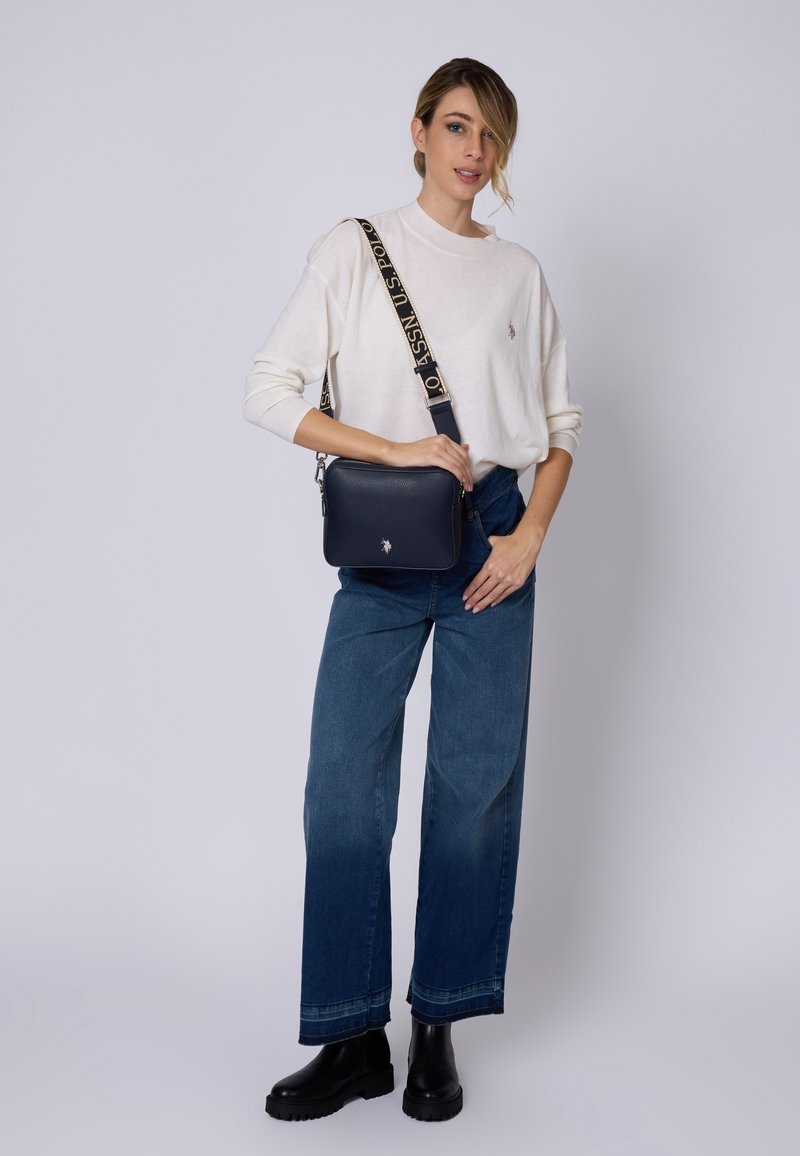 Námorně modrá kožená crossbody taška s texturovaným povrchem a vzorovaným popruhem. Modelka má na sobě bílé svetr a modré volné džíny. Černé boty.