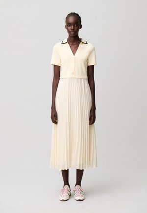 Mujer que lleva una blusa cremosa de manga corta con botones y ribete negro, combinada con una falda midi plisada a juego y zapatillas blancas con cordones rosas.