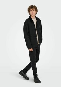 Ung mand med krøllet hår iført en sort jakke, beige sweater, sorte bukser og sorte klodsede sko, stående med det ene ben foran.