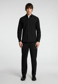 Camicia nera con bottoni in una vestibilità slim, con maniche lunghe, colletto classico e una texture liscia, abbinata a pantaloni e scarpe neri.