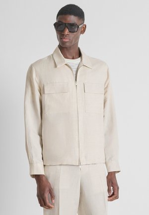 Homme portant une veste beige zippée avec poches poitrine, pantalon assorti et grandes lunettes de soleil noires sur fond uni.