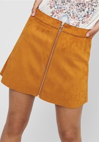 Suede mini kjol i senapsgul med en frontdragkedja och cirkulär draghandtag. Utsvängd design som vidgar sig något vid fållen.