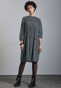Vestido geométrico gris con mangas largas, cintura fruncida y silueta relajada, combinado con botas negras hasta el tobillo.