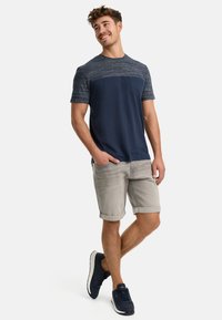 Marineblaues und graues Kurzarmhemd mit einer Tasche, kombiniert mit hochgekrempelten grauen Shorts und dunklen Turnschuhen, präsentiert ein lässiges Outfit.