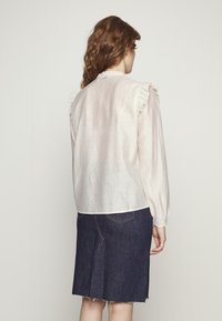 Blusa beige chiaro con dettagli a volant sulle spalle, maniche lunghe e tessuto testurizzato, abbinata a una gonna di jeans scura con orlo sfrangiato.