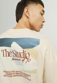 Beige oversized t-shirt met meerkleurige abstracte vormen en tekst "The Studio summer of 90" op de achterkant. Gemaakt van katoen.