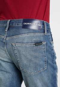 Blå denimjeans med en blekt finish, med en marinblå läderpatch vid midjan och signaturstickning på bakfickorna.