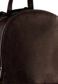 Bruine leren rugtas met een gestructureerd oppervlak, ronde vorm en zilveren ritsdetail aan één kant. Duidelijke stiksels langs de randen.