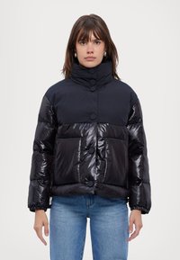Giacca a vento nera con colletto alto, texture lucida, grandi bottoni frontali e due tasche a toppa sul davanti, abbinata a jeans azzurri chiari.