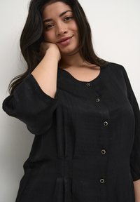 Blusa negra texturizada con tres botones que presentan herrajes dorados, escote amplio y mangas hasta los codos. Tejido suave con un brillo sutil.
