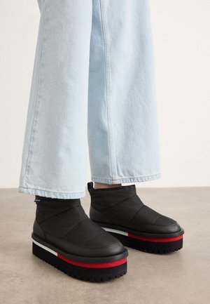 Czarne botki na platformie z czerwono-białym paskiem, noszone z jasnoniebieskimi szerokimi jeansami na beżowej podłodze.