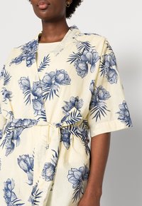 Vaaleankeltainen kimono-mallinen mekko, jossa on sinisiä kukkakuoseja, lyhyet hihat ja sidottava vyö. Valmistettu pehmeästä, teksturoidusta kankaasta.