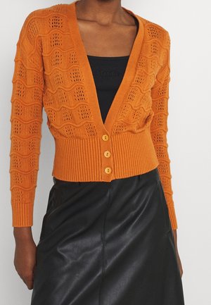 Femme portant un cardigan en maille orange avec des motifs ondulés, boutonné sur le devant, par-dessus un haut noir et une jupe en cuir noire.