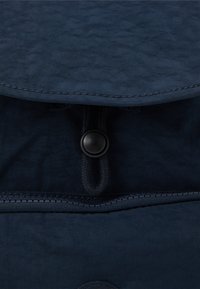 Laatikkomainen pimeansininen kangaspussi, jossa on teksturoitu pinta. Pussissa on nappisuljenta ja vetoketju alareunassa, joka takaa turvallisen pääsyn.