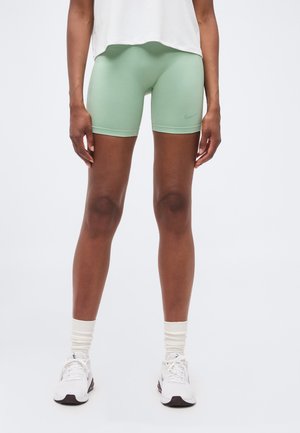 Osoba nosząca jasnozielone sportowe spodenki rowerowe, białą koszulkę, białe skarpetki i białe buty do biegania, stojąca na jednolitym tle.