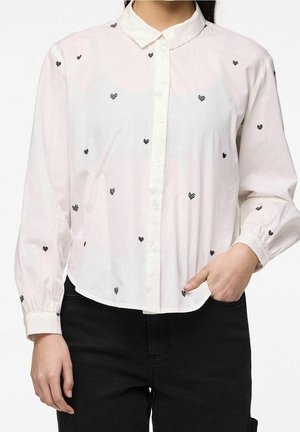 Kvinde iført en hvid skjorte med knapper og sorte pixelhjerte-mønstre samt sorte bukser, den ene hånd i lommen, foran en ensfarvet baggrund.